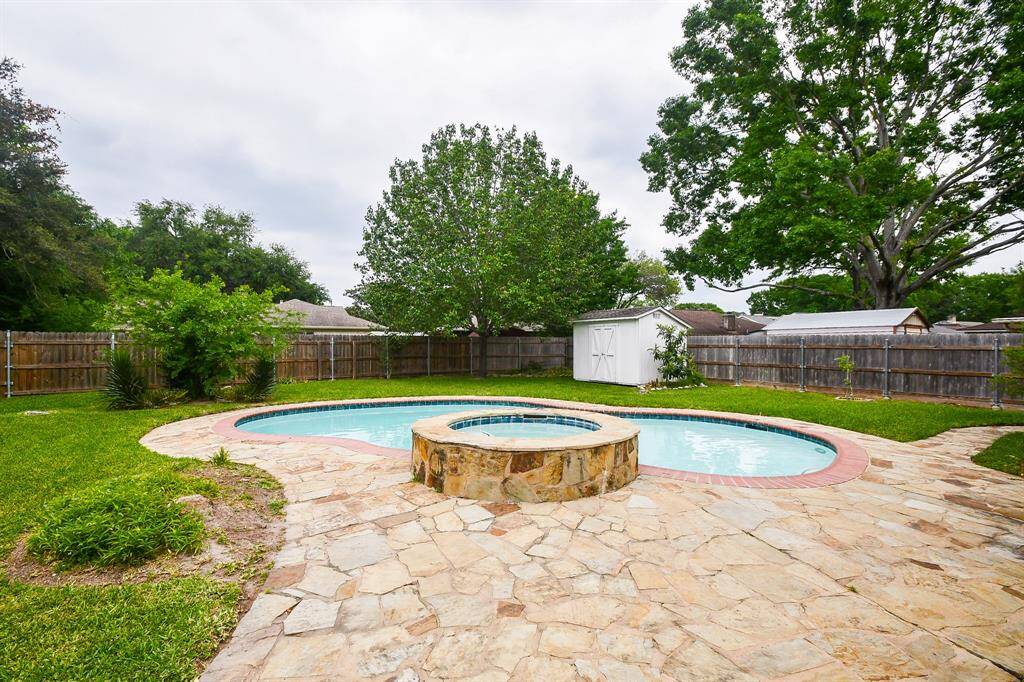 This backyard features a charming pool with a circular spa, surrounded by a stone patio. It's enclosed by a wooden fence with mature trees and a small storage shed, offering privacy and a serene atmosphere.