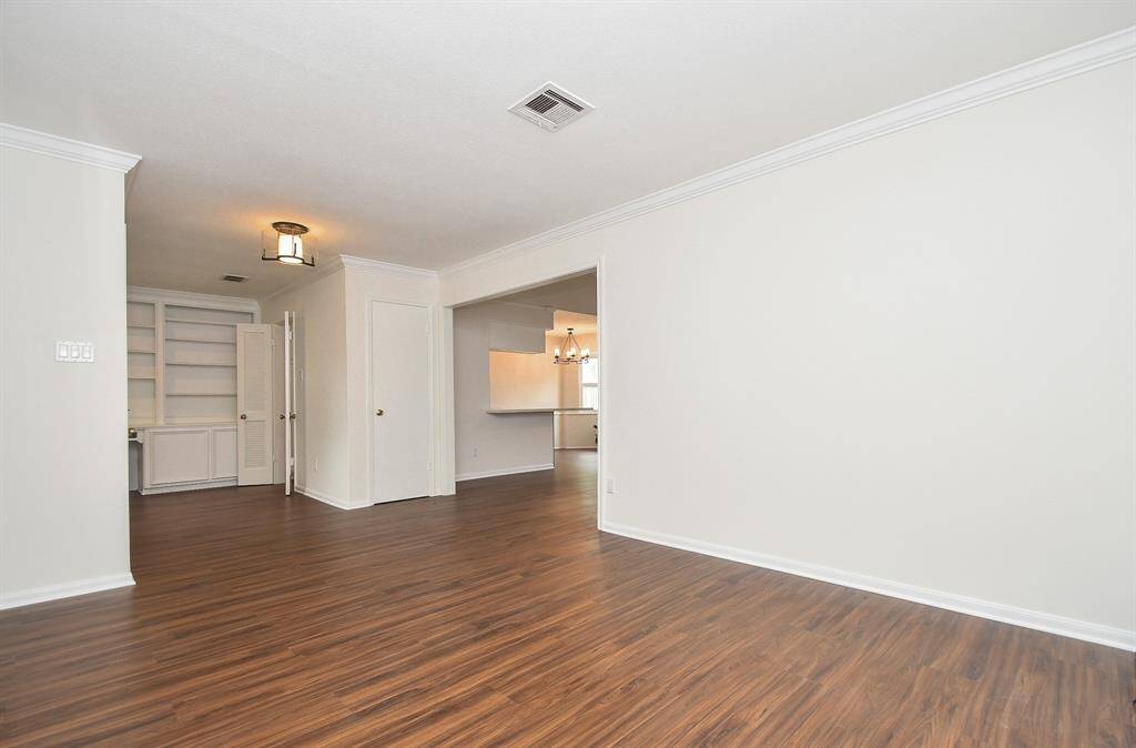 This photo showcases a spacious, light-filled room with wood look floors, neutral walls, and crown molding. It features built-in shelving, and an open layout leading to a kitchen area, perfect for versatile living and entertaining.