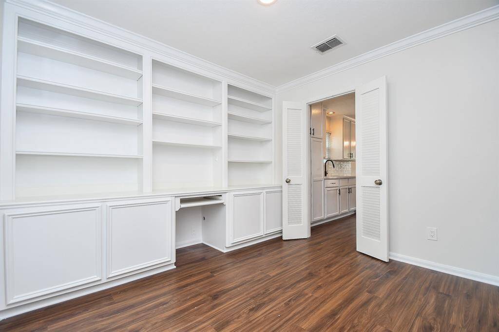 This room features built-in white shelving and cabinets, offering ample storage space. The wood look flooring adds warmth, and a doorway leads to a bright kitchen, enhancing the home's flow and functionality.