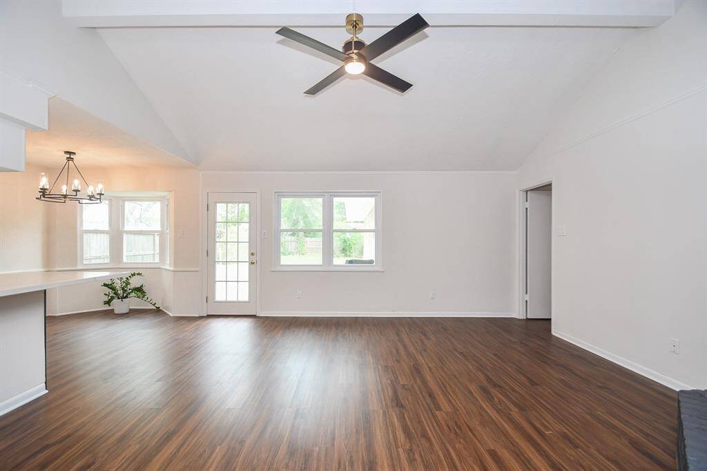 This spacious living area features vaulted ceilings with a modern ceiling fan, hardwood floors, and ample natural light from large windows and a glass door leading outside. The open layout connects to a cozy dining nook with a stylish chandelier.