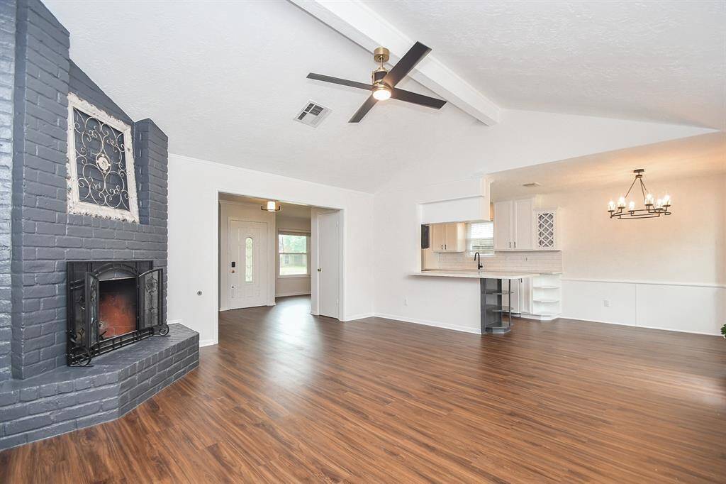 This spacious living area features elegant wood look flooring, a stylish brick fireplace, and a modern led ceiling fan. The open layout seamlessly connects to a bright kitchen and dining space, enhanced by a chic chandelier and ample natural light.