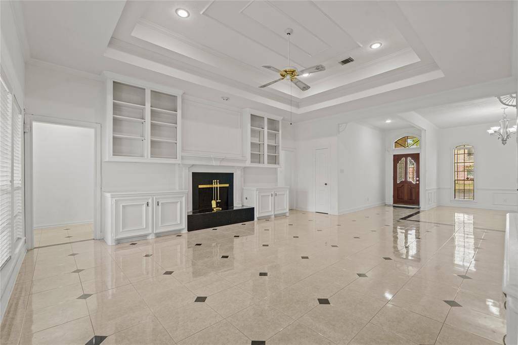 This stunning living area is the perfect blend of elegance and warmth, designed to impress while feeling like home. The soaring tray ceiling with recessed lighting adds a touch of grandeur, while the gleaming tile floors reflect natural light beautifully, making the space feel even more open and inviting. A sophisticated fireplace, framed by built-in shelving, creates the ideal focal point for cozy nights in or entertaining guests. The seamless flow into the foyer, with its charming arched windows