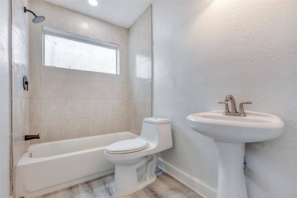 This bathroom features a modern design with a tiled shower and bathtub combination, a sleek pedestal sink, and a contemporary toilet. Natural light filters through a frosted window, enhancing the clean, fresh look.  Mirror to be installed.