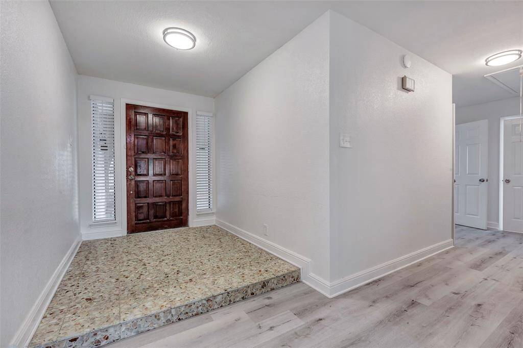 Inviting entryway with a rich wooden front door, flanked by two narrow windows with blinds. The space features a small step-up with terrazzo flooring leading to a bright, open hallway with light-colored laminate flooring and modern lighting.