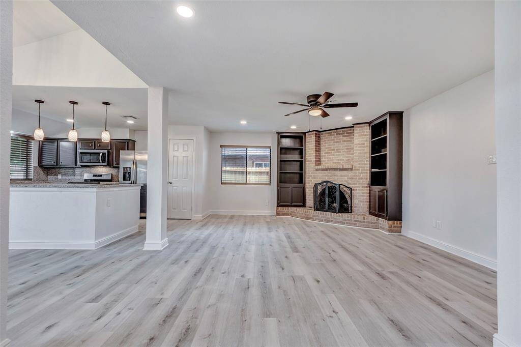 This spacious room features modern laminate flooring, a cozy brick fireplace with built-in shelving, and a ceiling fan. The open layout leads to a contemporary kitchen with pendant lighting and stainless steel appliances, perfect for entertaining.