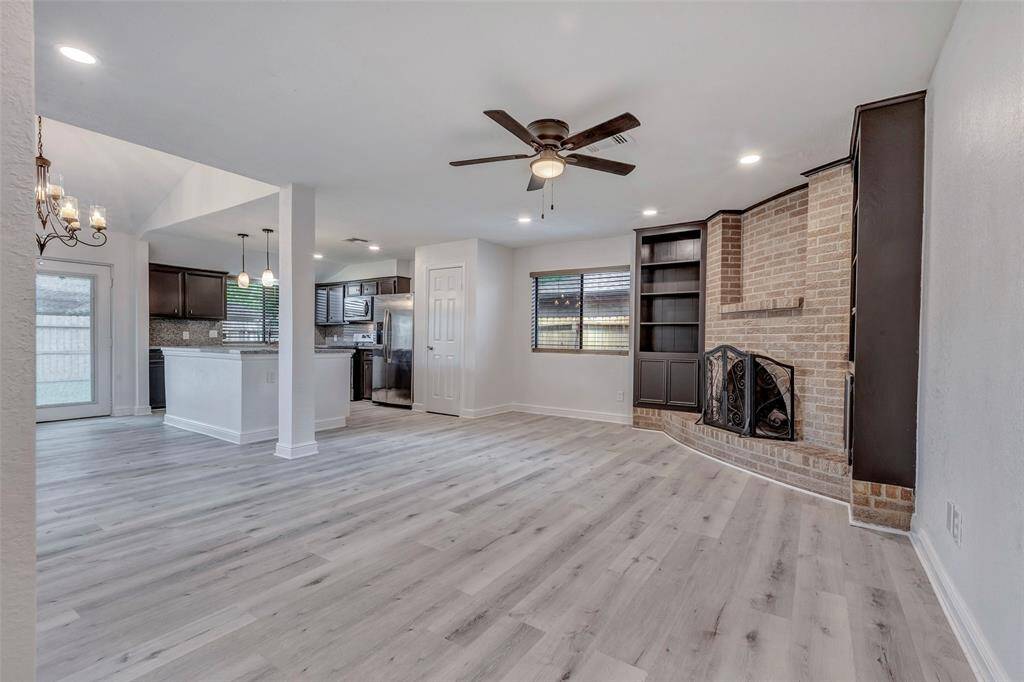 This living area features modern light wood flooring, a brick fireplace with built-in shelving, and an open layout connecting to a stylish kitchen with dark cabinetry and stainless steel appliances. The room is bright with recessed lighting, a ceiling fan, and a chandelier over the dining area, creating a cozy and inviting atmosphere.