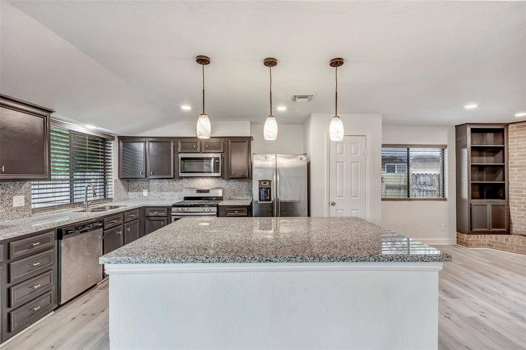 This photo showcases a modern kitchen with a large granite island, dark cabinetry, and stainless steel appliances. It features pendant lighting and an open layout, leading to a cozy living area with a built-in shelf and brick accent.