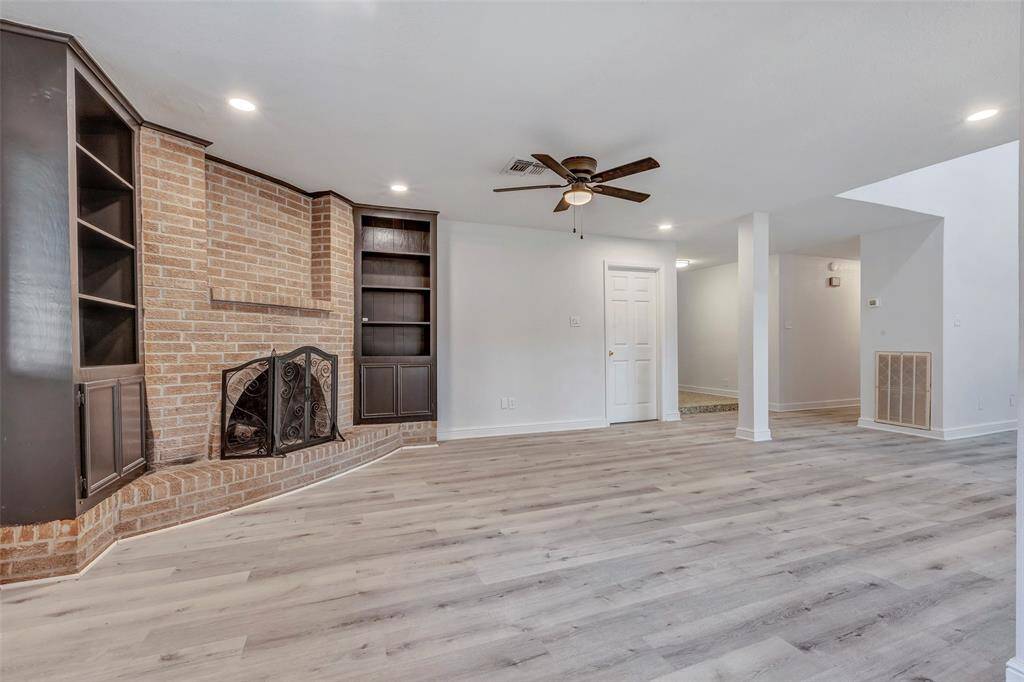 This spacious living room features a cozy brick fireplace with built-in shelves, modern light wood look laminate  flooring, and a ceiling fan. The room is bright and open, perfect for family gatherings.
