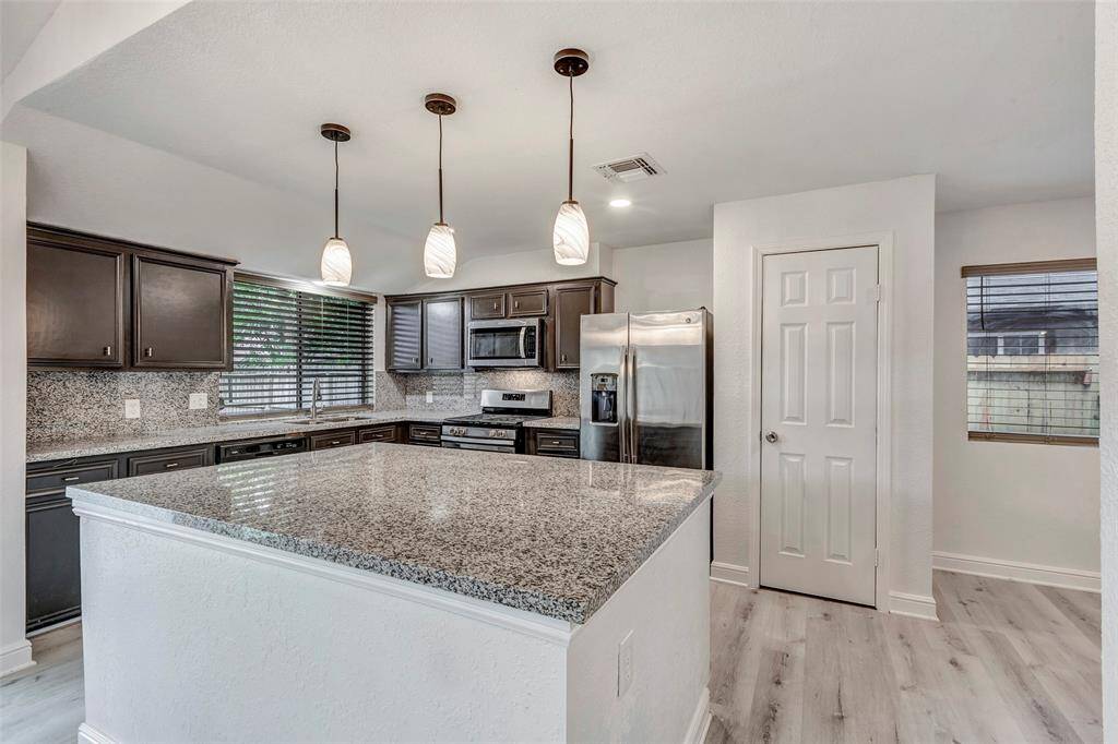 This kitchen features modern stainless steel appliances, granite countertops, and dark cabinetry. It includes a large central island with pendant lighting and wood-style flooring.