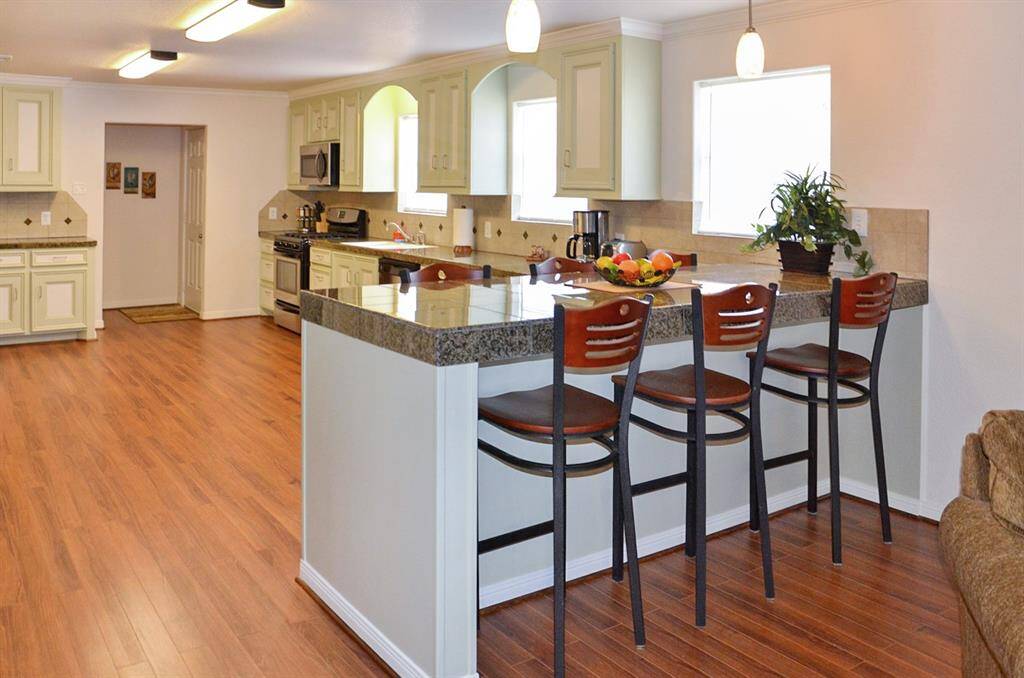 A spacious, open-concept living area with wood flooring flows into a bright kitchen featuring a breakfast bar and modern appliances. The room is warmly decorated with comfortable seating and stylish wall art, offering a welcoming atmosphere.