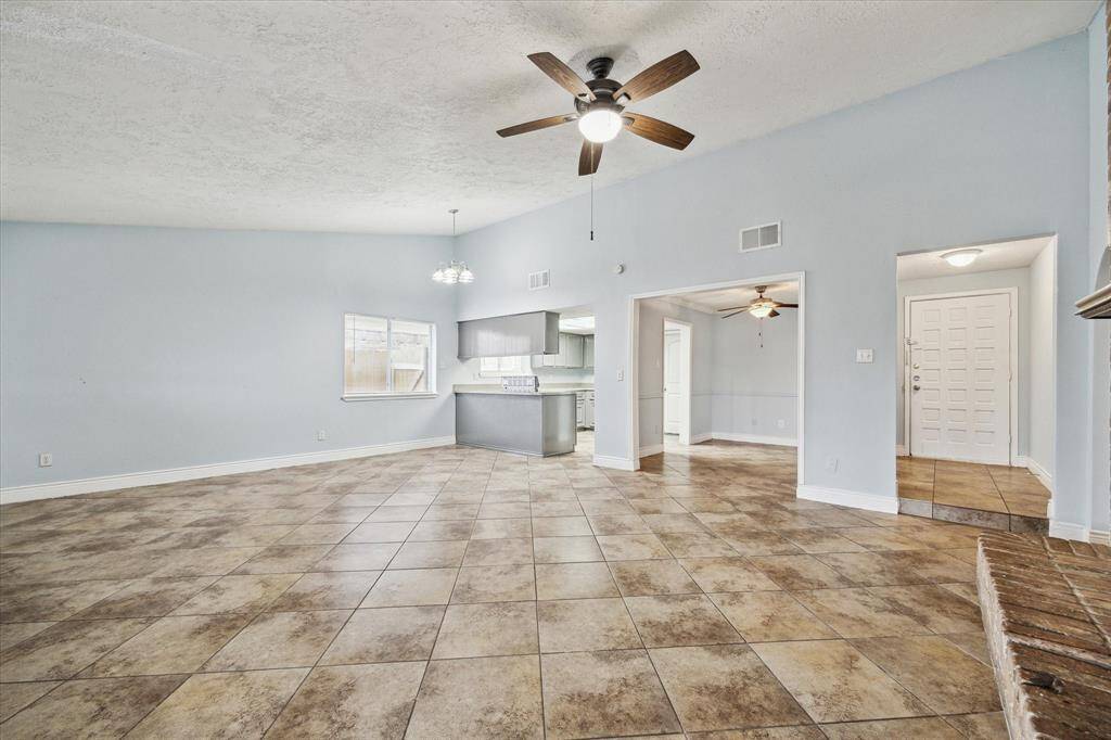 This spacious living area features tile flooring, light blue walls, and a ceiling fan. It's open to the kitchen and has ample natural light through several windows. The layout includes easy access to adjacent rooms and the entryway.