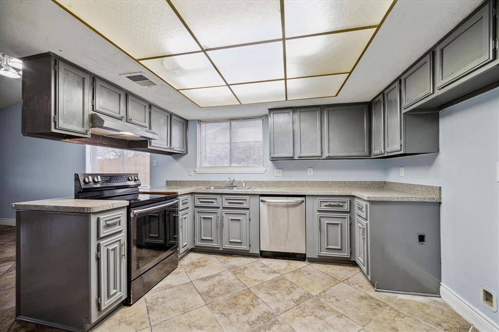 This kitchen features a newer stove with vent hood and a dishwasher . The space is well-lit with a large window and overhead lighting, complemented by neutral-toned countertops and tile flooring.