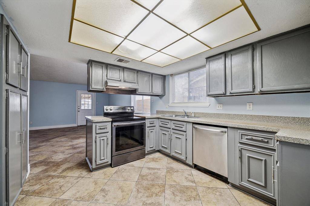This kitchen features a newer stove with vent hood and a dishwasher . The space is well-lit with a large window and overhead lighting, complemented by neutral-toned countertops and tile flooring.