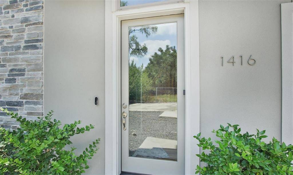 Modern entrance featuring a full-heightglass door with side panels that allowample natural light, flanked bystonework and green shrubbery.