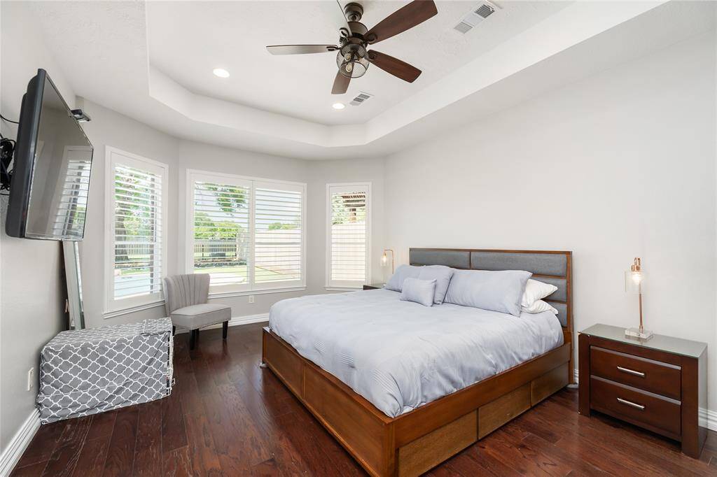 This serene primary bedroom offers a spacious layout enhanced by rich hardwood floors and an elegant tray ceiling with a ceiling fan. Large windows with plantation shutters frame peaceful views and fill the room with natural light, creating a warm and restful atmosphere perfect for unwinding at the end of the day.