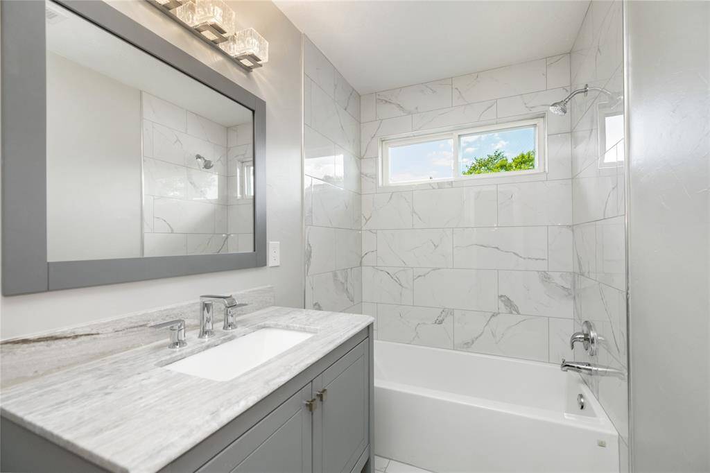 This elegantly designed secondary bathroom showcases marble-inspired tiles that add a touch of luxury, paired with a modern vanity and sleek countertop. A large mirror enhances the spacious feel, while a beautifully framed window invites natural light, brightening the bathtub area and creating a serene, spa-like atmosphere.