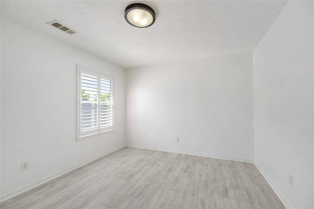 This airy room features light wood flooring, soft white walls, and large windows with classic shutters, allowing natural light to flood the space. A modern ceiling light adds a touch of elegance, creating a versatile area perfect for a guest room, home office, or additional living space.