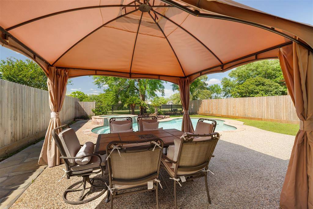 This inviting backyard features a covered patio, complete with a charming table and chairs, overlooking a sparkling swimming pool. Surrounded by lush greenery and enclosed by a wooden fence, the space offers a private and tranquil setting—perfect for relaxing, dining, or entertaining outdoors.