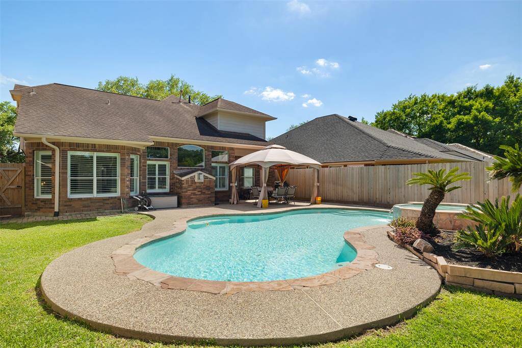 This stunning backyard features a gracefully curved pool with elegant stone edging, complemented by a manicured lawn that enhances the serene setting. A shaded patio area with comfortable seating offers the perfect spot for relaxation or entertaining, while the surrounding wooden fence ensures privacy and tranquility.