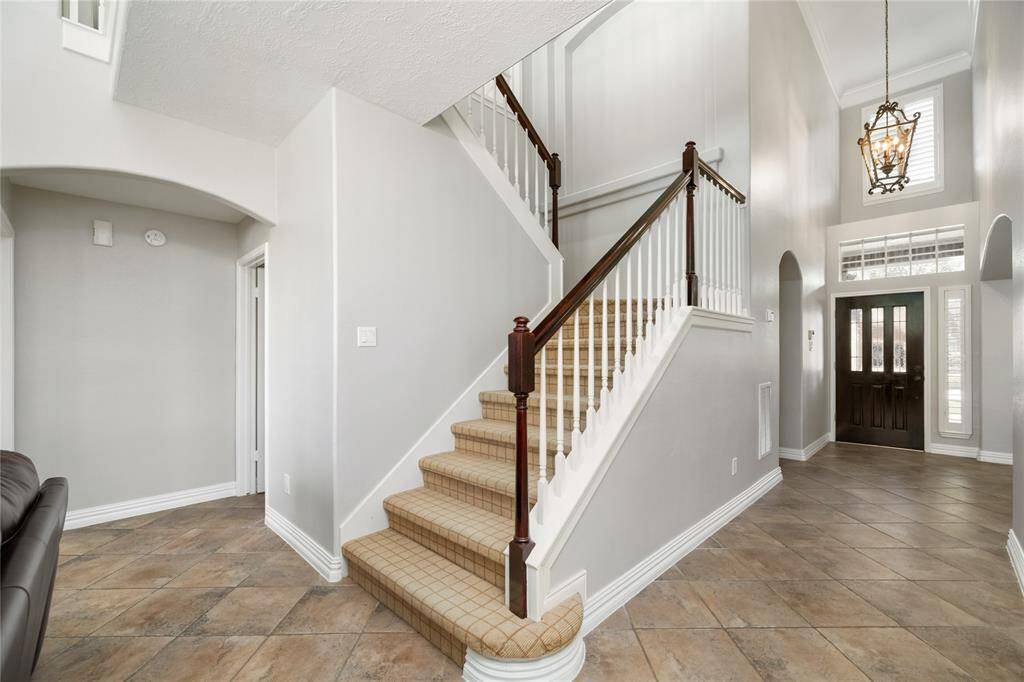 A soaring ceiling and graceful chandelier set the tone in this elegant foyer, where neutral tones and classic tile flooring create a warm, airy ambiance. The staircase, adorned with rich wood accents and plush carpeting, offers a sophisticated focal point, while the dark wood entry door adds a touch of contrast and charm.