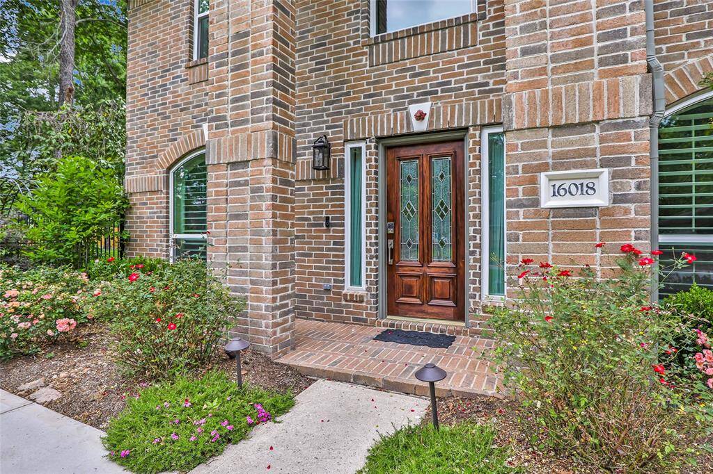 This photo showcases a charming brick home with a pitched roof, surrounded by a lush green lawn and mature trees. The property features a fenced yard and a spacious driveway leading to a double garage, offering both privacy and curb appeal.