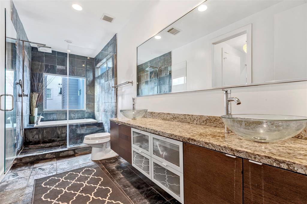 This spacious main bedroom en-suite featuring dual glass washbasins, a tub and shower combo, large vanity mirrors, and sleek dark floor tiles for a luxurious touch.