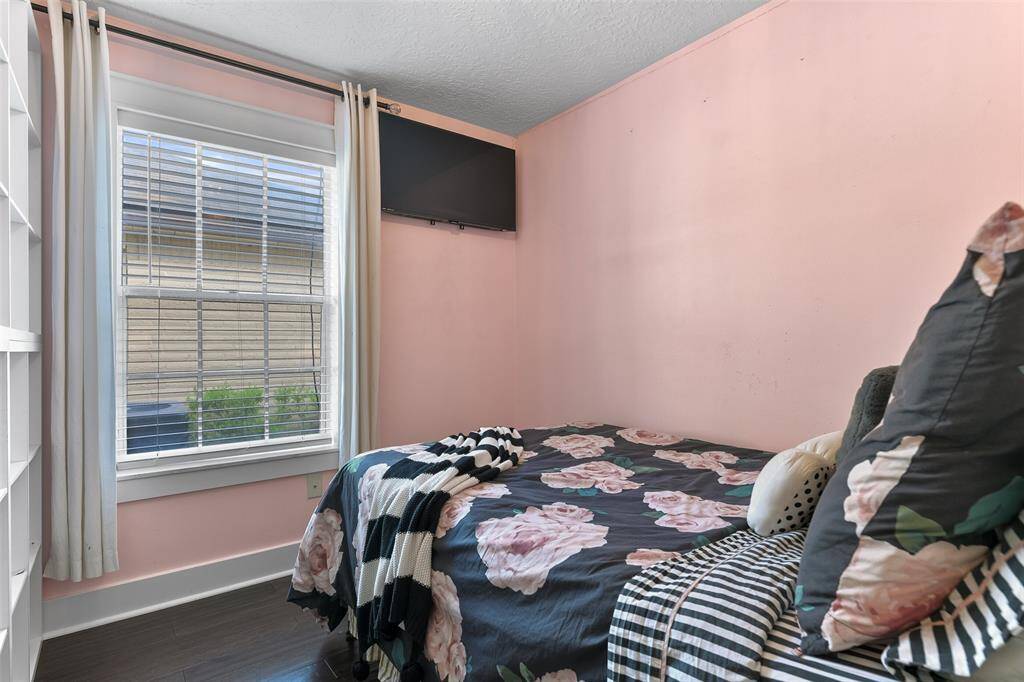 The first of three secondary bedrooms in the home features a rose pink paint selection, a delicate chandelier, dark wood flooring, a side-facing window with 2-inch blinds, and a closet with built-in shelves.