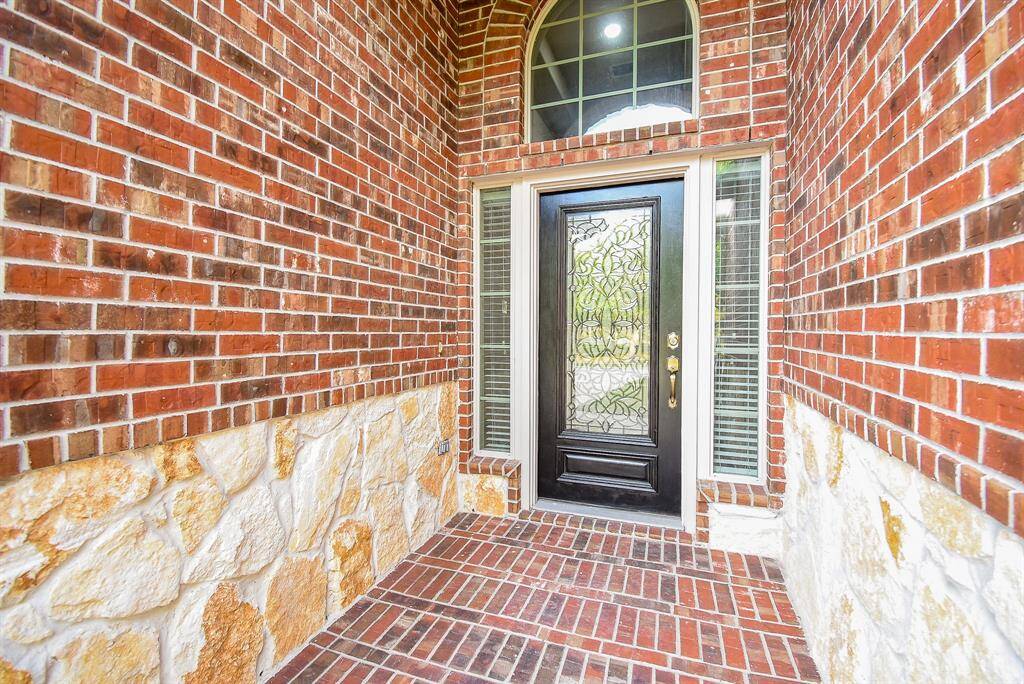 This entryway features a welcoming black door with intricate glass design, framed by sidelights and an arched transom window. The surrounding walls are a mix of red brick and natural stone, creating a warm, inviting atmosphere.