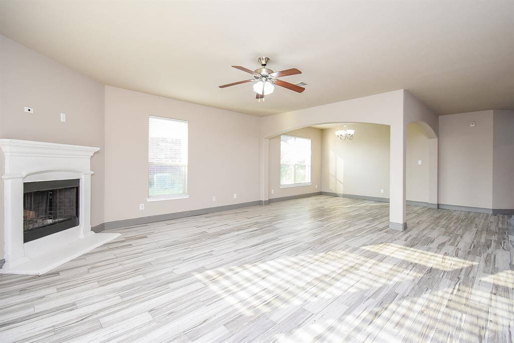 This spacious living area features elegant wood-style flooring, a cozy fireplace, and abundant natural light. It seamlessly connects to a dining space with a chandelier, creating an inviting and open atmosphere perfect for entertaining.