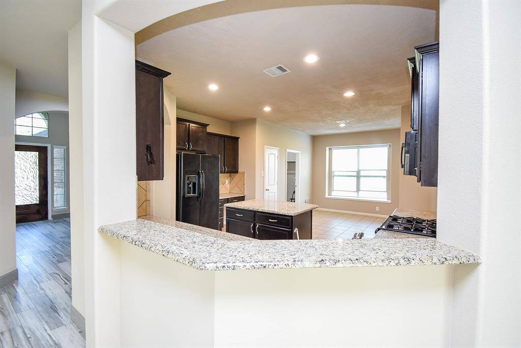 This photo showcases a modern kitchen with granite countertops, dark cabinetry, and a center island. It features stainless steel appliances, recessed lighting, and a large window for natural light. The open layout connects to a hallway with wood-look flooring, leading to the entrance.