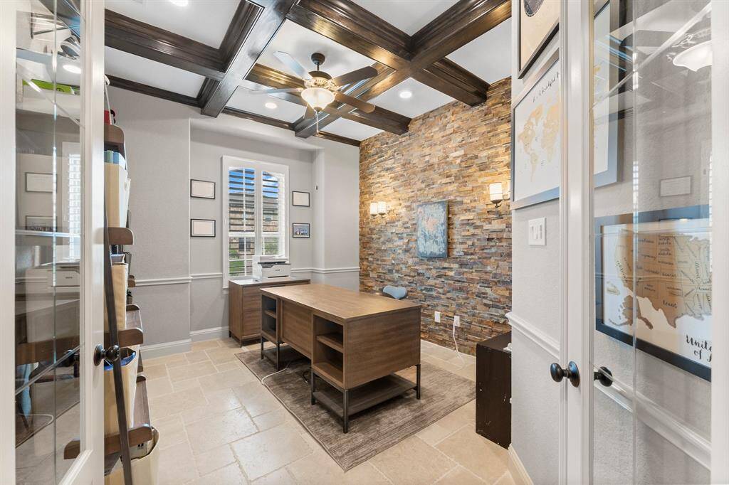 This photo showcases a stylish home office with a stone accent wall, coffered ceiling with wood beams, and a modern ceiling fan. The room features a large window with shutters, built-in shelving, creating a sophisticated and functional workspace.