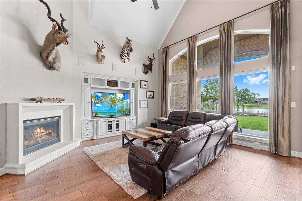 Spacious living room with high ceilings, large windows offering natural light and views of the greenery outside. Features a cozy fireplace, hardwood floors, and a built-in entertainment center.