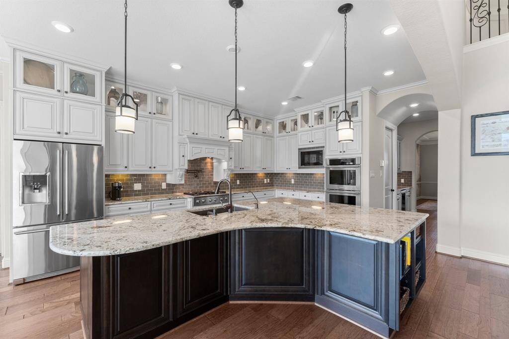 This kitchen features a spacious layout with a large granite island, stainless steel appliances, and elegant pendant lighting. It has white cabinetry with glass display sections and a stylish backsplash, ideal for modern living.