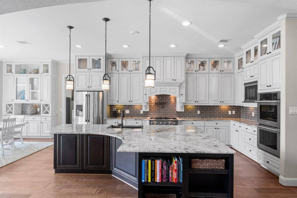 This modern kitchen features elegant white cabinetry with glass accents, a spacious granite island with built-in shelving, and stainless steel appliances. It includes pendant lighting, a tiled backsplash, and an open layout with a dining area, perfect for entertaining and family gatherings.