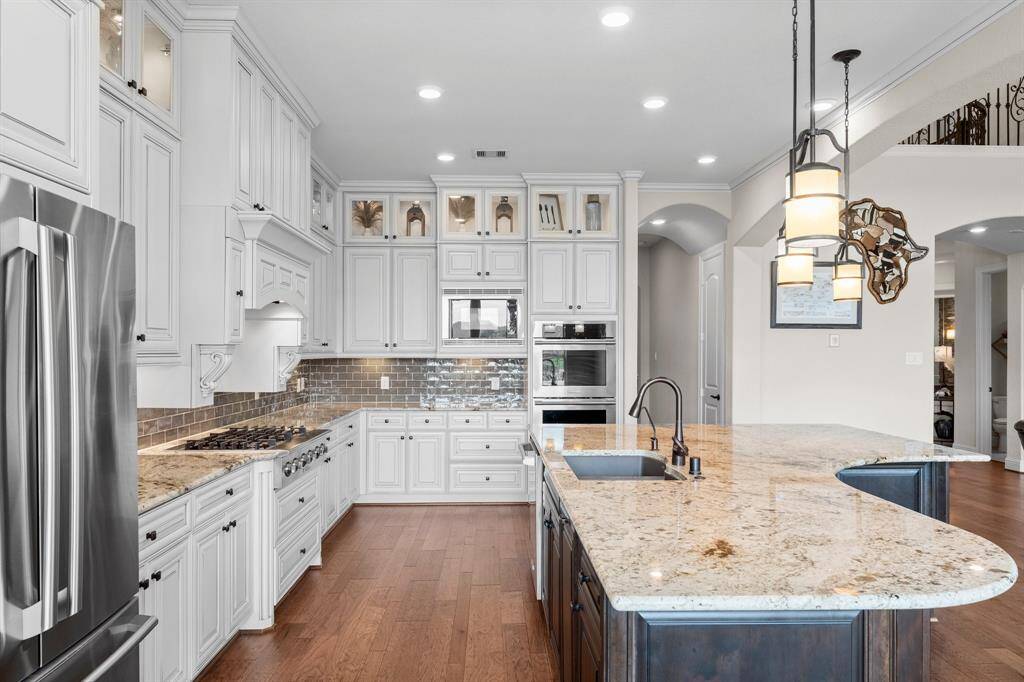 This kitchen features elegant white cabinetry, stainless steel appliances, and a spacious granite island with a built-in sink. The hardwood floors and modern lighting create a warm and inviting atmosphere, perfect for cooking and entertaining.