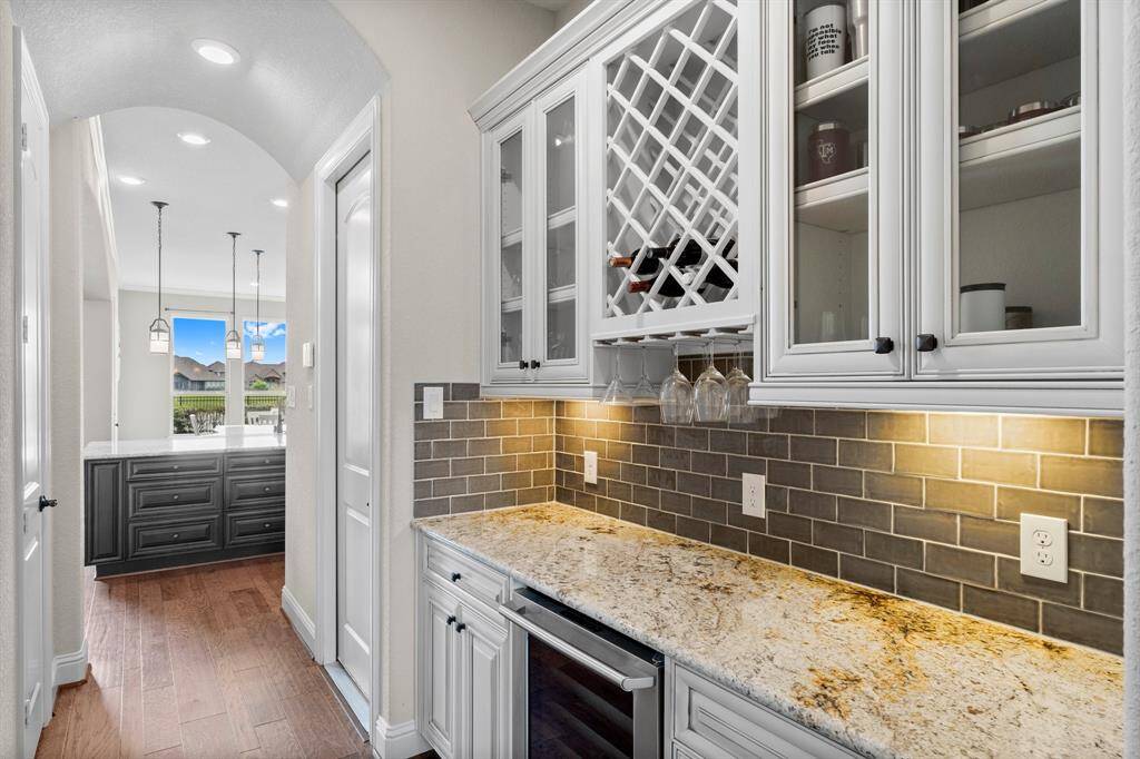 This photo showcases a stylish butler's pantry with granite countertops, glass-front cabinetry, and a wine rack. It features a subway tile backsplash and elegant under-cabinet lighting. The space connects to a bright room with pendant lights and a view of the outdoors.
