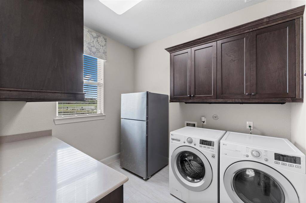 This photo shows a well-lit laundry room featuring a washer and dryer, ample dark wood cabinetry for storage, a countertop for folding, and a window providing natural light.