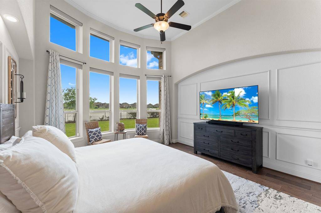 This spacious bedroom features large, curved windows offering ample natural light and a serene view. It includes a ceiling fan, elegant wainscoting, and hardwood floors, creating a cozy, inviting atmosphere.