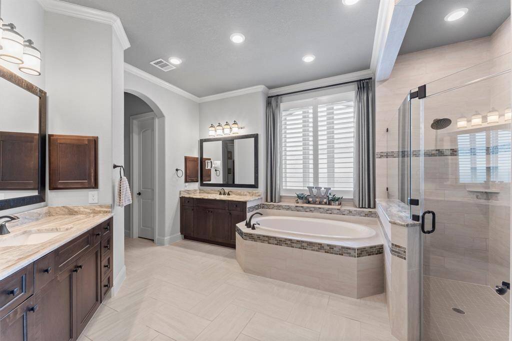 This spacious bathroom features elegant tile flooring, a large soaking tub with a tiled surround, and a walk-in glass shower. Dual vanities with stylish dark wood cabinetry offer ample storage. Recessed lighting and large windows provide a bright, inviting atmosphere.