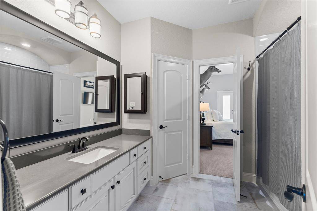 This bathroom features a modern design with a sleek gray countertop and ample cabinet space. It includes a large mirror, stylish lighting fixtures, and a shower with a curtain. The room connects to a bedroom, visible through the open door.