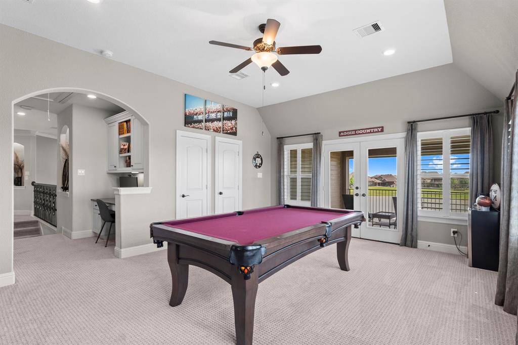This spacious game room features a pool table, carpeted flooring, and a ceiling fan. It includes a built-in desk area and offers plenty of natural light through French doors leading to a balcony with scenic views.