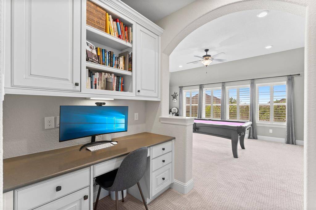 This photo showcases a bright and inviting home office space with built-in shelves and a desk, leading into a spacious recreation room with large windows, a pool table, and ceiling fan, perfect for work and leisure.