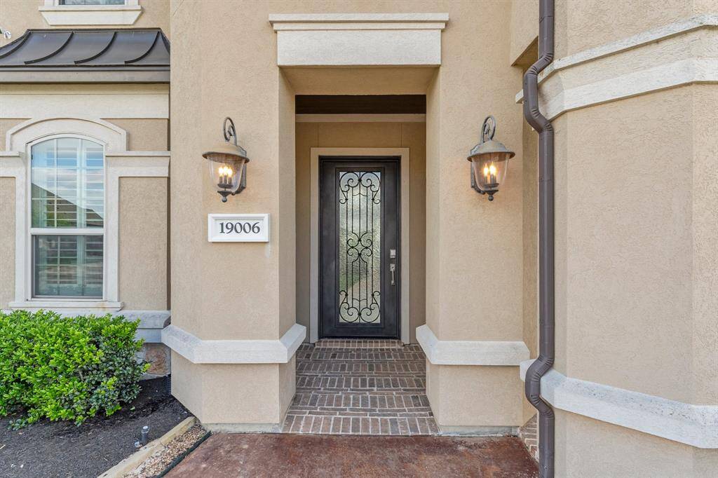 This photo showcases an elegant entryway with a decorative wrought iron door, flanked by stylish lantern-style lights. The exterior features warm beige tones and white trim, with a neatly landscaped area including greenery.