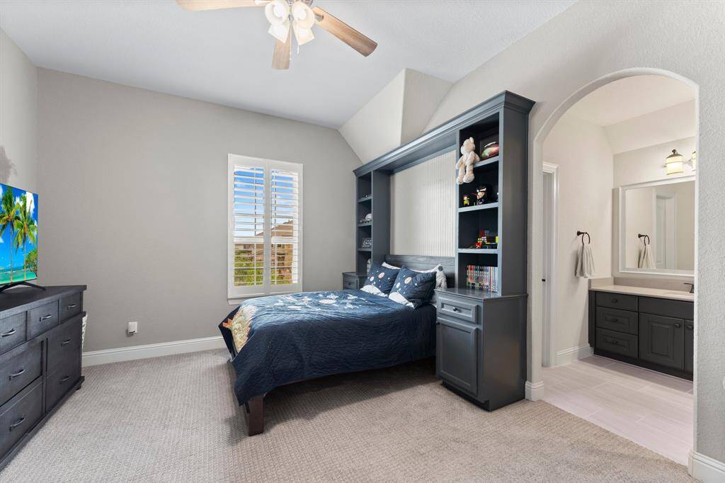 This bedroom features a cozy carpeted floor, and a ceiling fan. Natural light streams through the window with plantation shutters. An en-suite bathroom is accessible through an arched entryway.