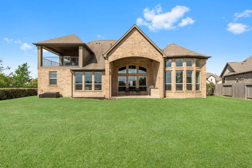 This photo shows the backyard of a spacious brick home featuring large arched windows and a covered patio. The expansive lawn offers plenty of outdoor space, and the home has a second-story balcony.