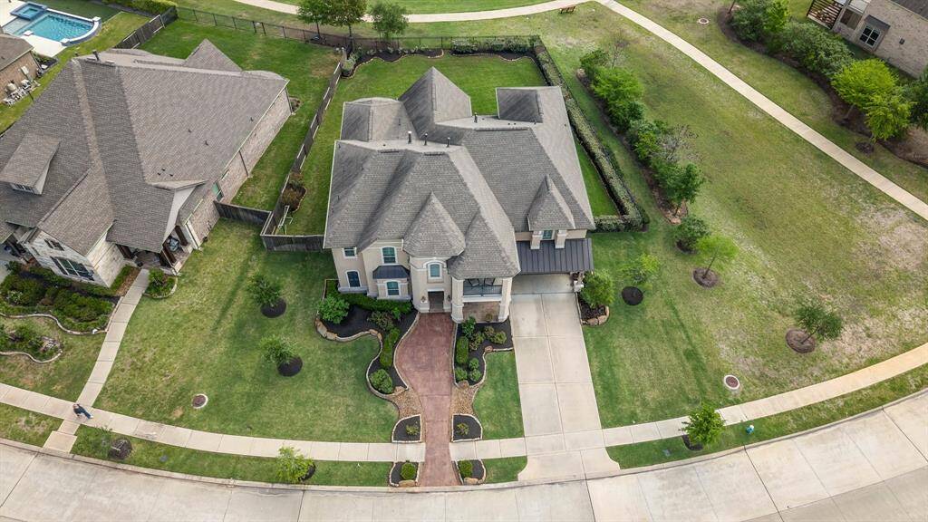 This aerial view showcases a spacious, elegant two-story home with a unique, multi-gabled roof design. It features a well-maintained front yard with landscaped pathways and ample driveway space. The property is situated in a serene neighborhood with nearby greenery, offering a sense of privacy and tranquility.