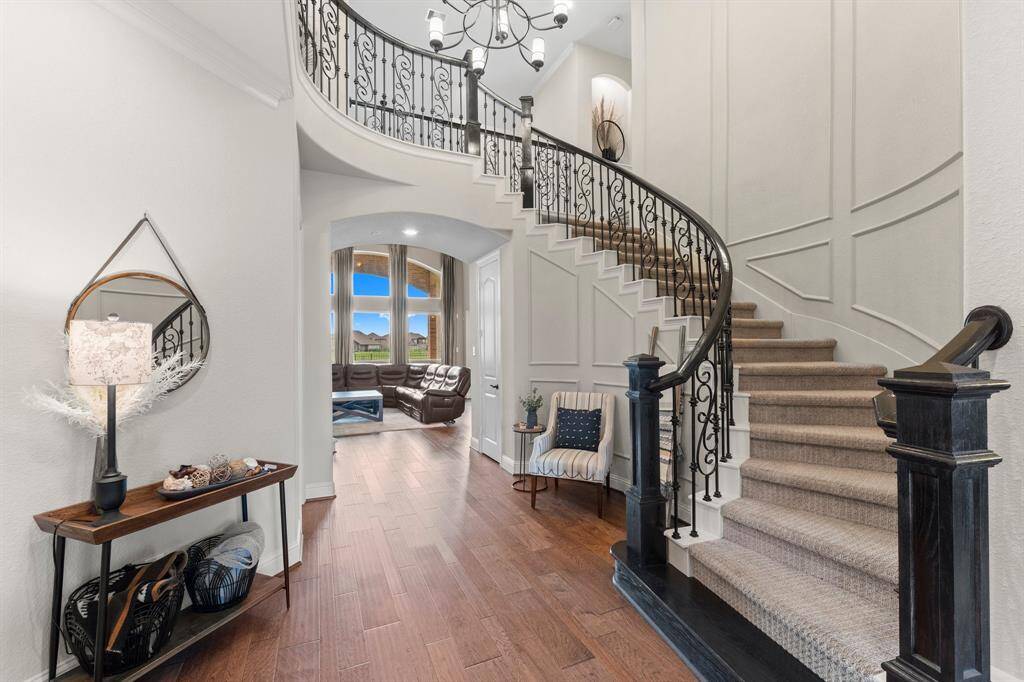 This elegant entryway features a grand curved staircase with plush carpeting and ornate iron railing. The space is accentuated by a stylish chandelier, rich wood flooring, and a cozy sitting area. An inviting glimpse into the living room showcases large windows and ample natural light.