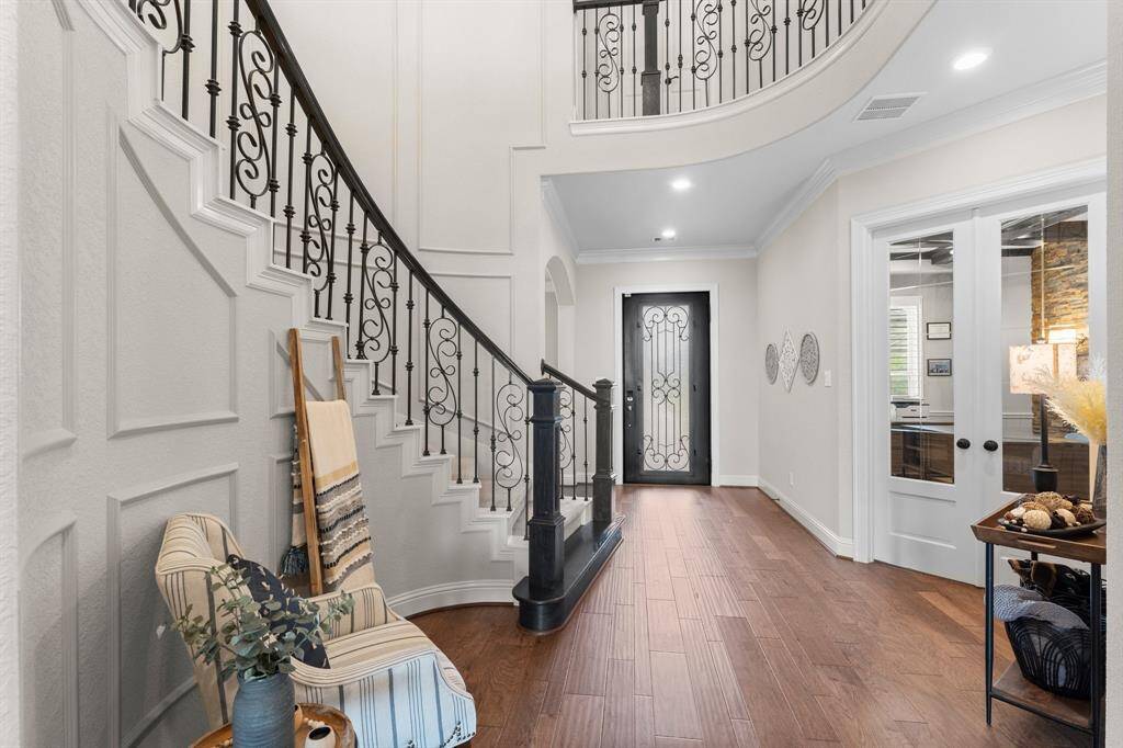 This inviting entryway features a grand curved staircase with elegant wrought iron railings, warm hardwood floors, and a stylish glass-panel door. The space is complemented by a cozy chair with decorative accents and leads to a room with double glass doors, creating an open and welcoming atmosphere.