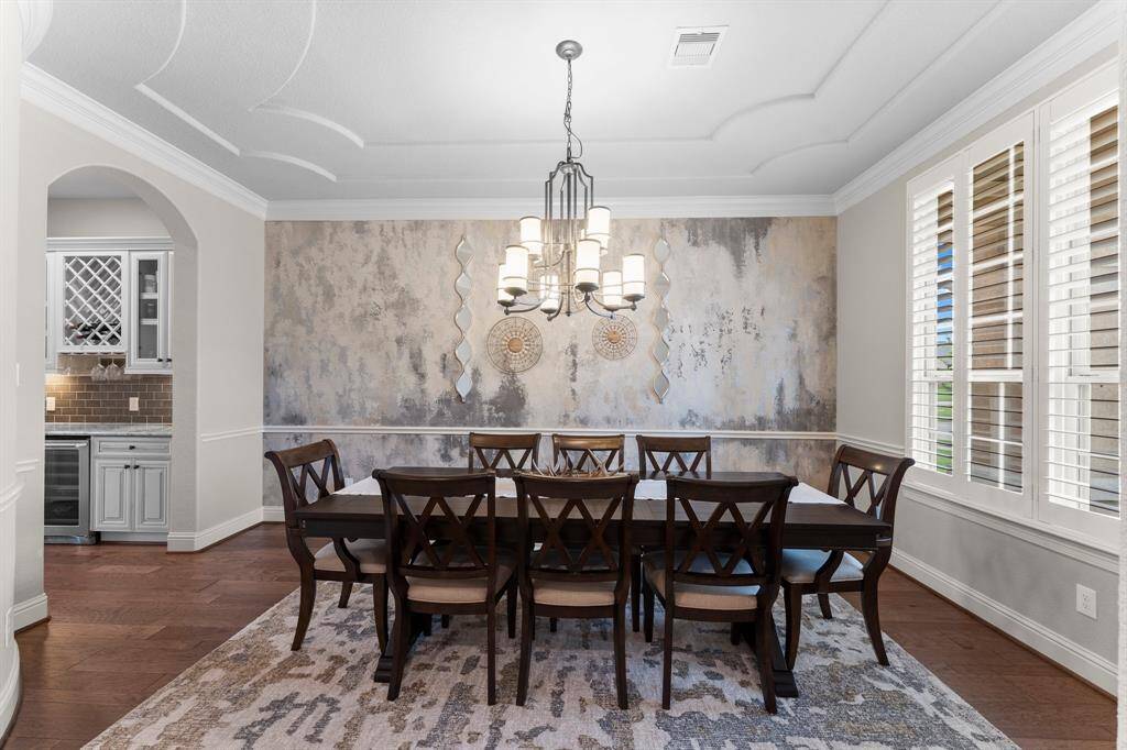 The photo showcases an elegant dining room with a stylish chandelier, textured accent wall, and hardwood floors. Natural light streams in through large windows with plantation shutters. An arched entry leads to a small adjoining bar area with cabinetry and a wine rack.