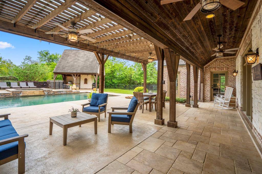 You will love this covered patio with rustic wood beams, ceiling fans, and room for seating. It overlooks a pool with a stone deck, surrounded by lush greenery and a charming pool house, perfect for relaxation and entertaining.